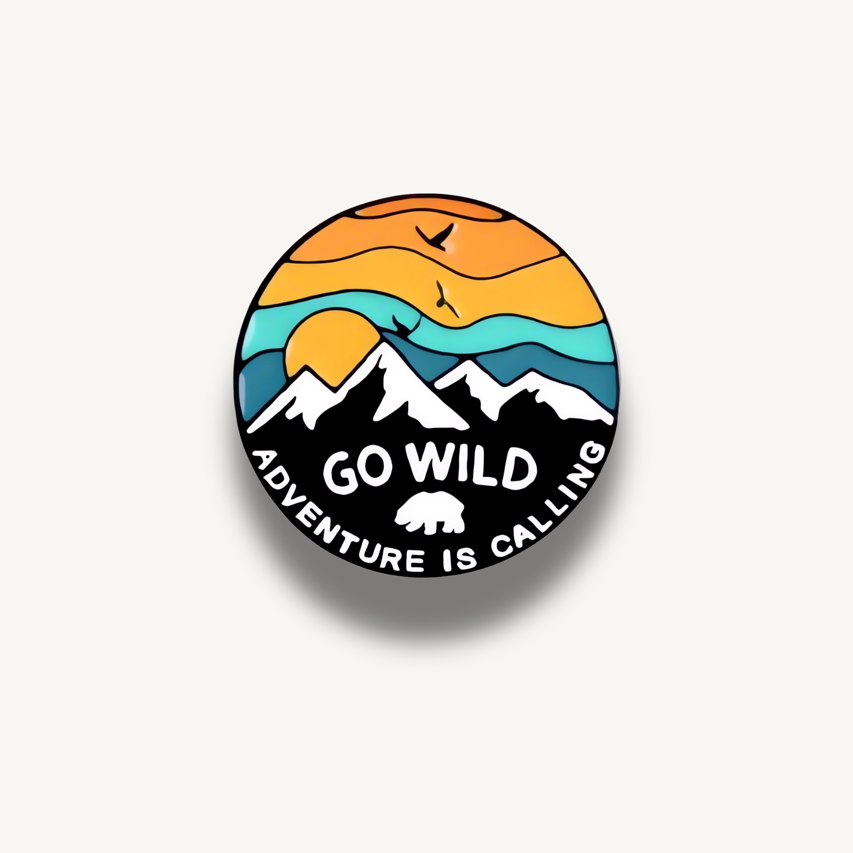 Kruhová brošňa s motívom hôr, západu slnka, lietajúcich vtákov a medveďa, s textom 'GO WILD ADVENTURE IS CALLING'.