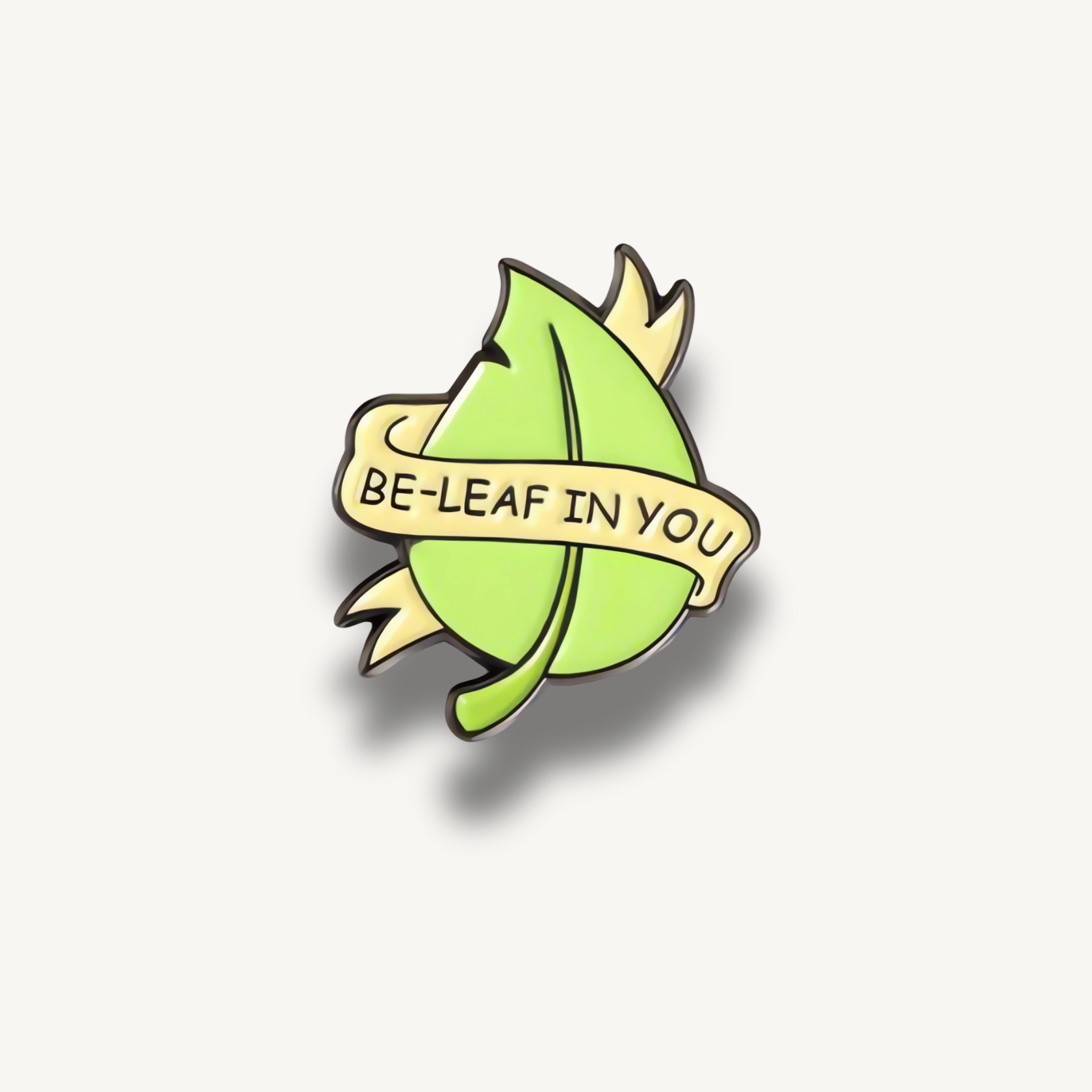 Smaltovaná brošňa v tvare svetlozeleného listu so žltou stuhou a čiernym nápisom 'BE-LEAF IN YOU'.