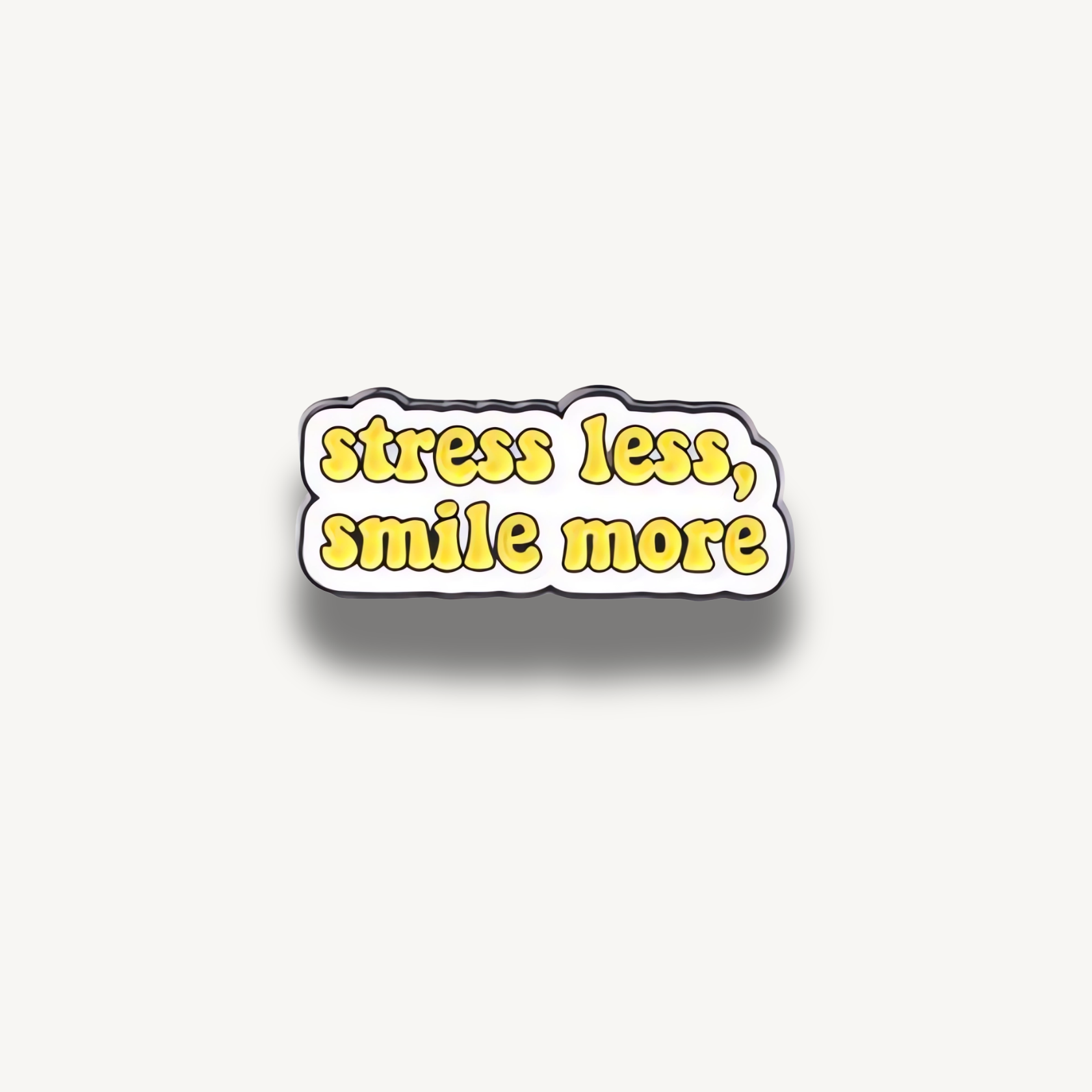 Smaltovaná brošňa s nápisom 'stress less, smile more' v žltom bublinkovom písme na bielom pozadí s čiernym okrajom.