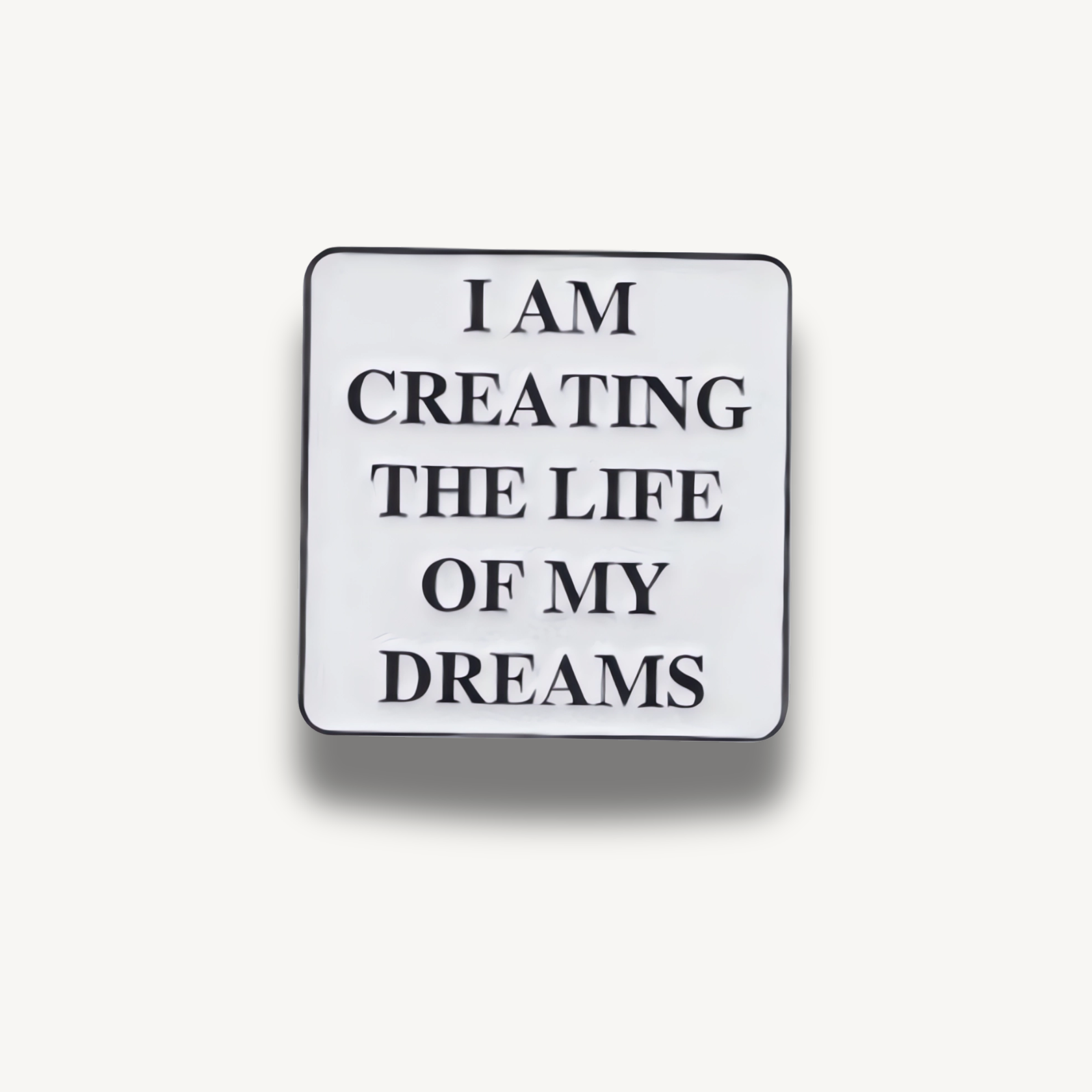 Biela štvorcová brošňa s čiernym nápisom 'I AM CREATING THE LIFE OF MY DREAMS'