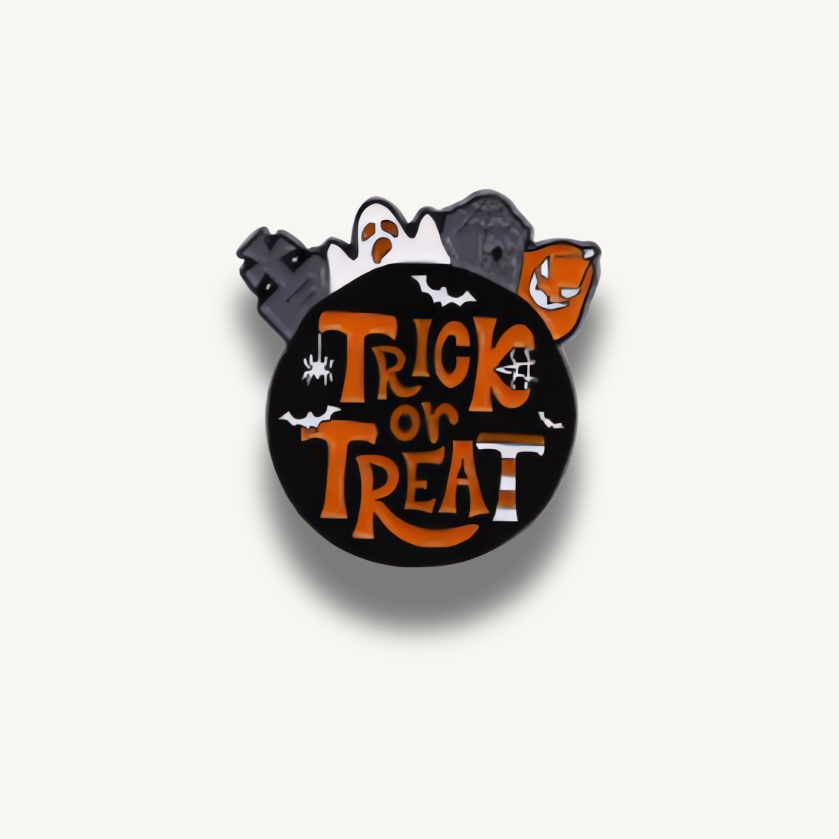 Okruhlá čierna brošňa s oranžovým nápisom Trick or Treat, tekvicou, duchom a náhrobnými kameňmi.