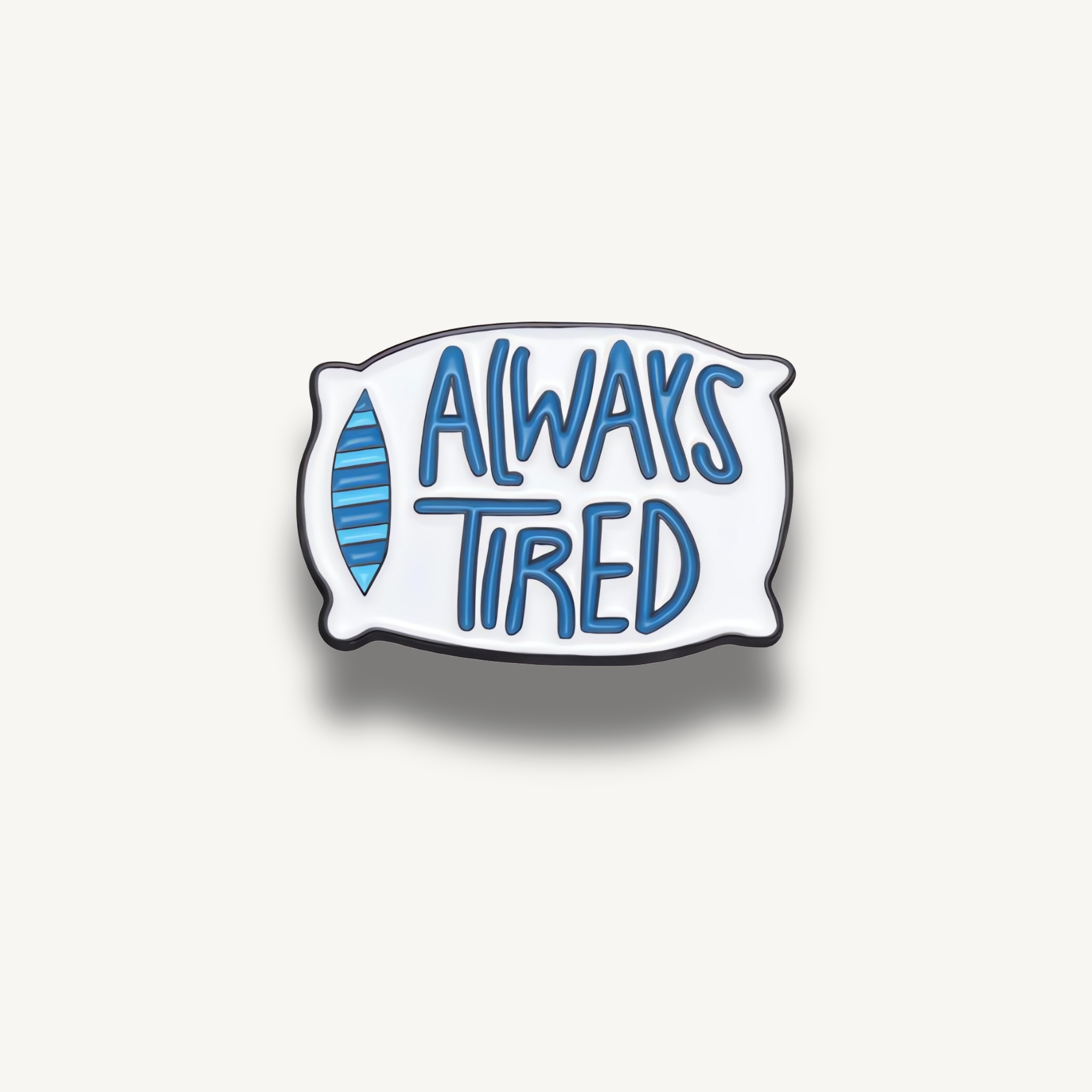 Smaltovaná brošňa v tvare bieleho vankúša s modrým nápisom Always Tired a pruhovanou maskou na spanie.