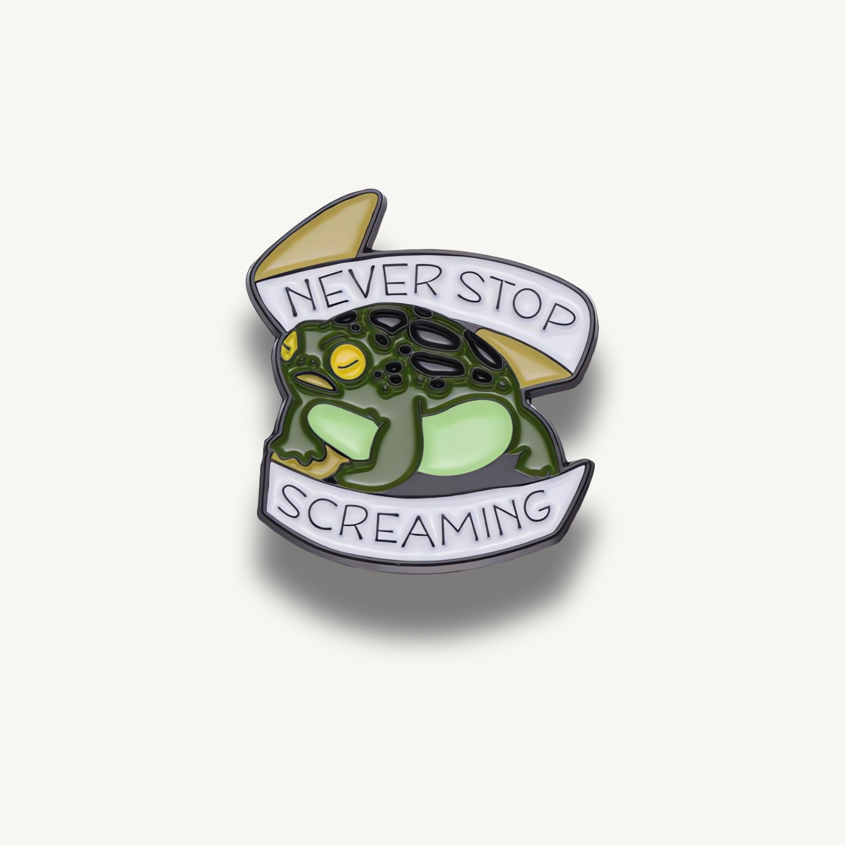 Smaltovaná brošňa zobrazujúca tmavozelenú žabu so žltými očami a bielym transparentom s textom 'NEVER STOP SCREAMING'.