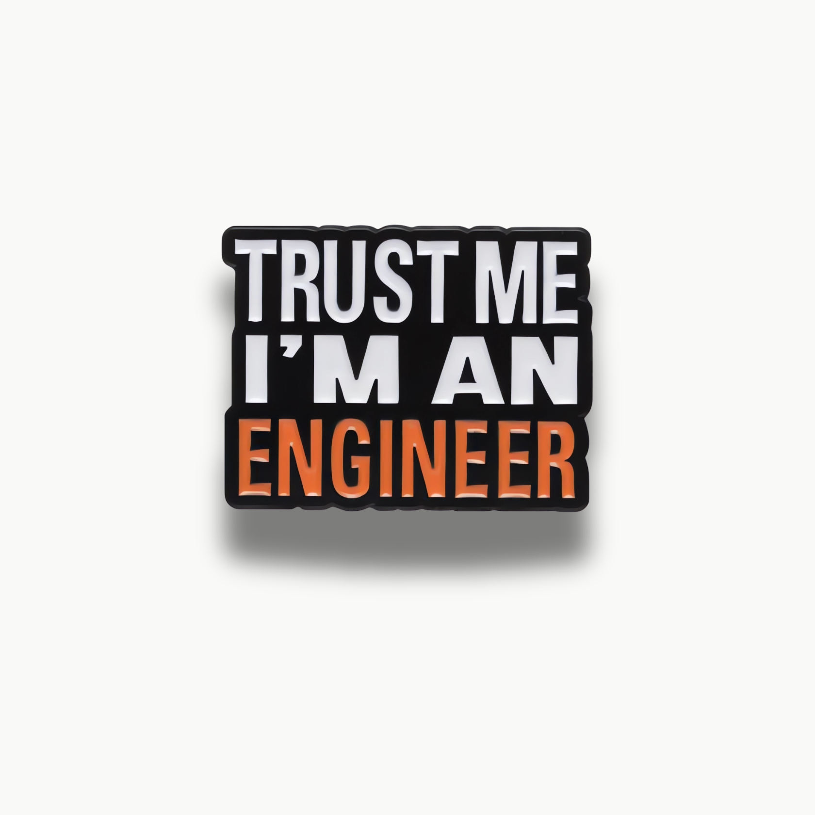 Čierna brošňa s nápisom 'TRUST ME I'M AN ENGINEER' v bielej a oranžovej farbe.