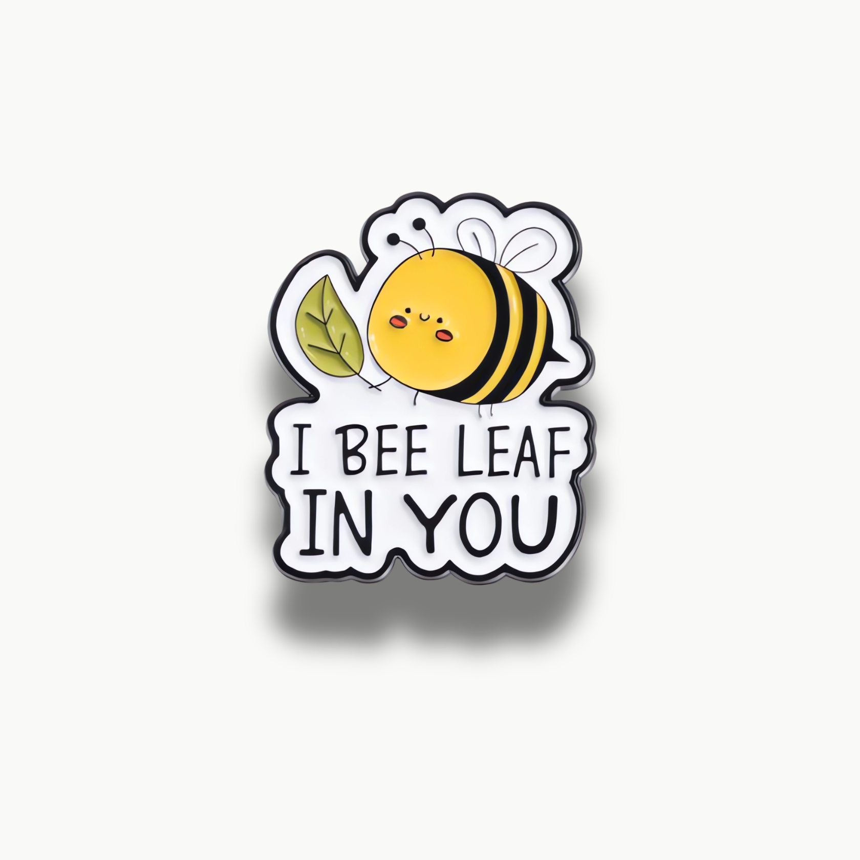 Smaltovaná brošňa s kreslenou žltou včielkou, zeleným listom a textom 'I Bee Leaf In You' na bielom podklade s čiernym okrajom.