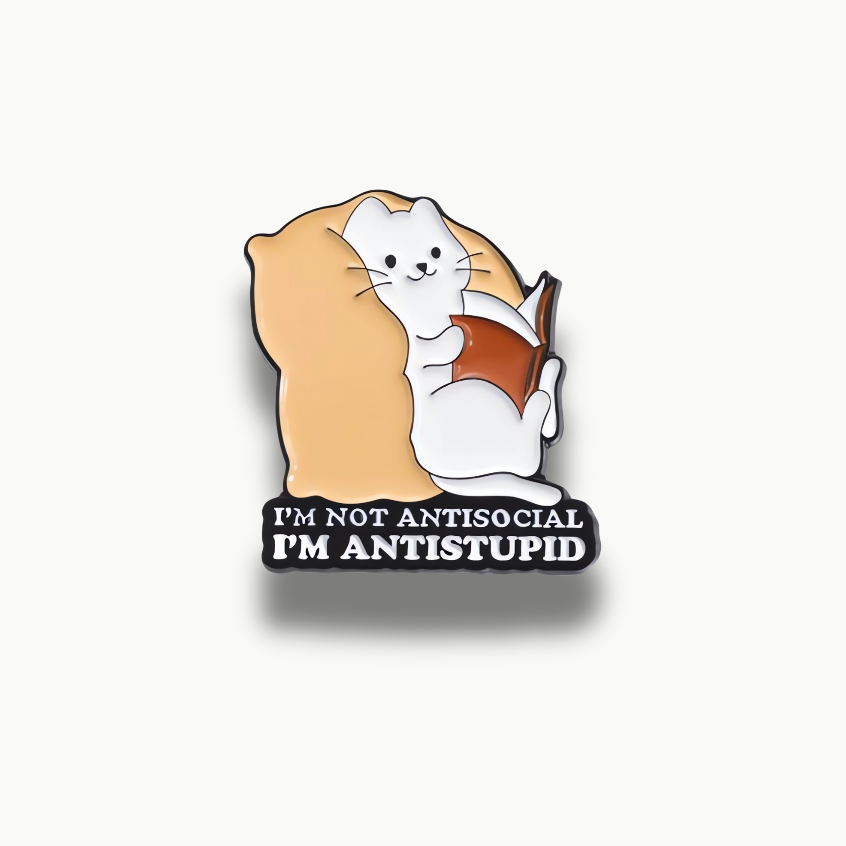 Smaltovaná brošňa biela mačka ležiaca na béžovom vankúši, čítajúca hnedú knihu, s textom 'I'M NOT ANTISOCIAL I'M ANTISTUPID'.