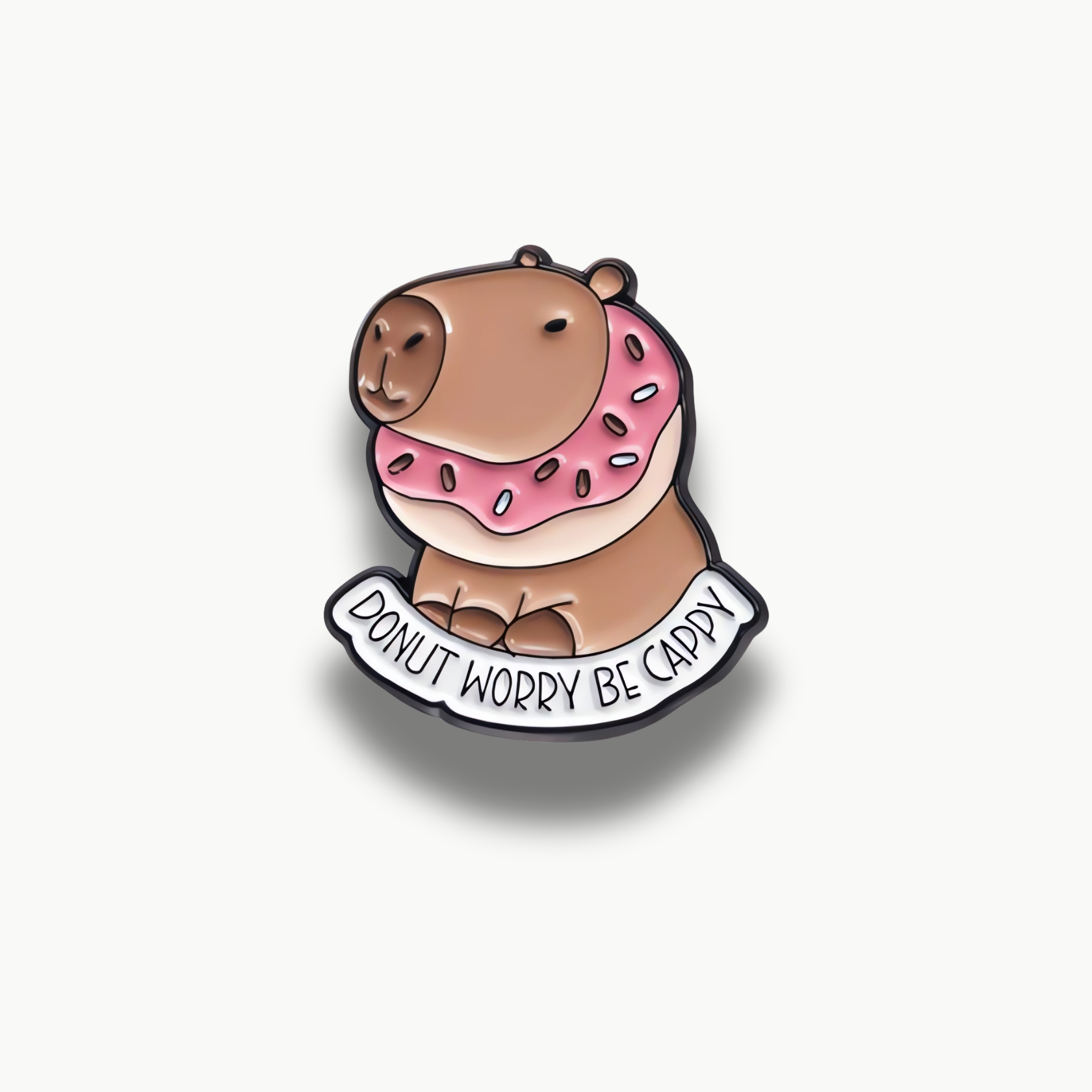 Smaltovaná brošňa zobrazujúca sediacu kapibaru s ružovou šiškou okolo krku a nápisom "DONUT WORRY BE CAPPY".