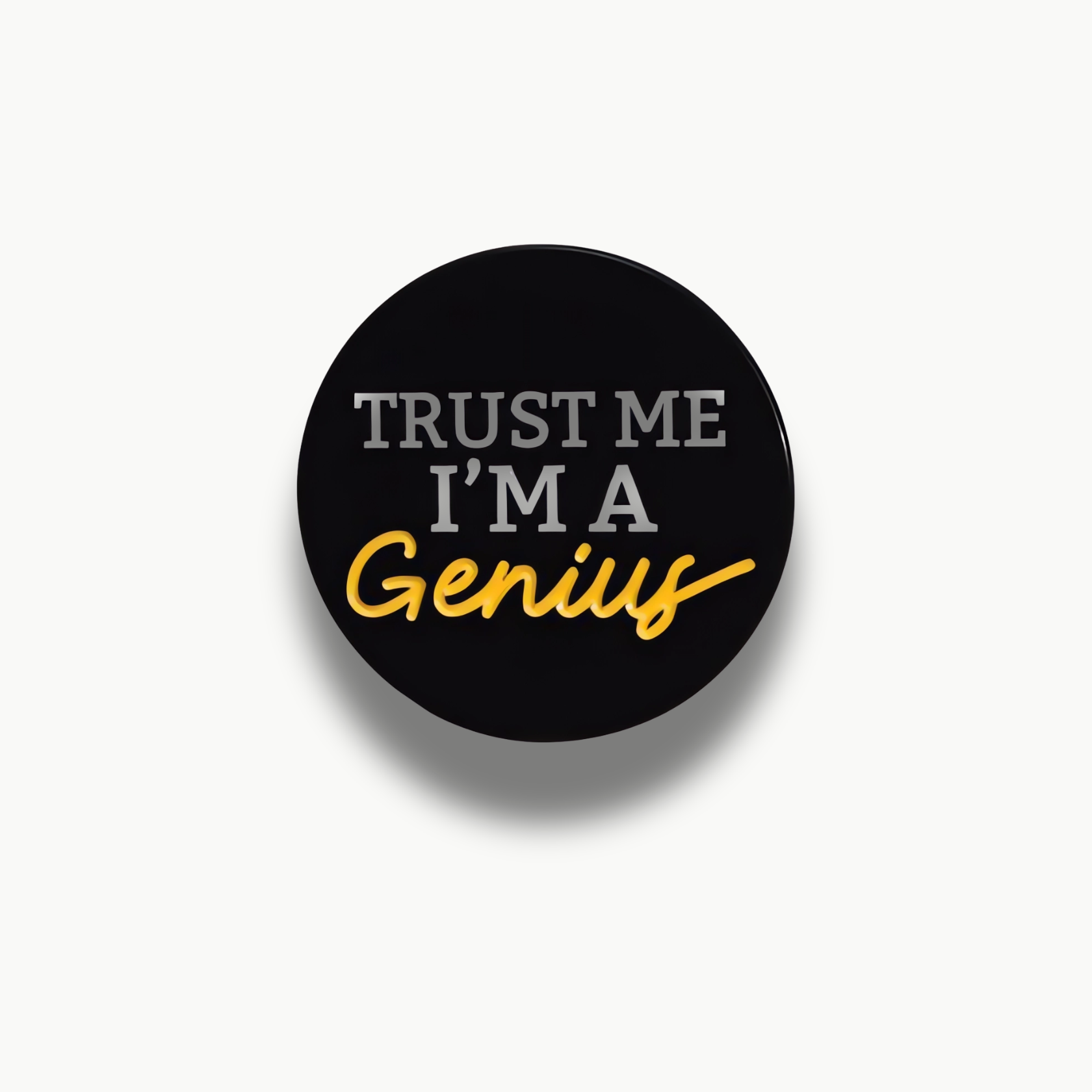 Čierna kruhová brošňa s bielym textom 'Trust Me I'm A' a žltým textom 'Genius'.