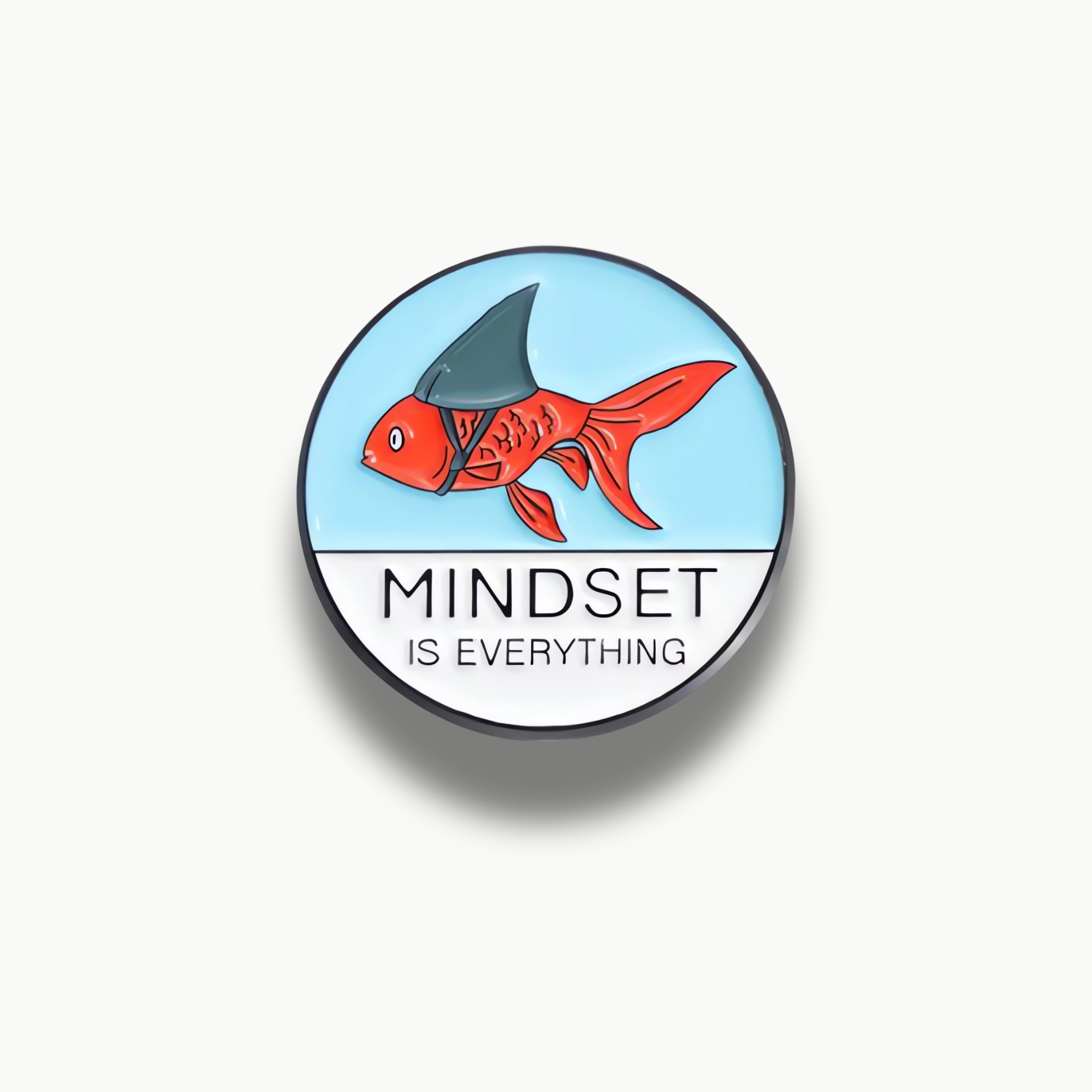 Okrúhla smaltovaná brošňa s oranžovou rybou, ktorá má na chrbte žraločiu plutvu a nápis 'MINDSET IS EVERYTHING'.