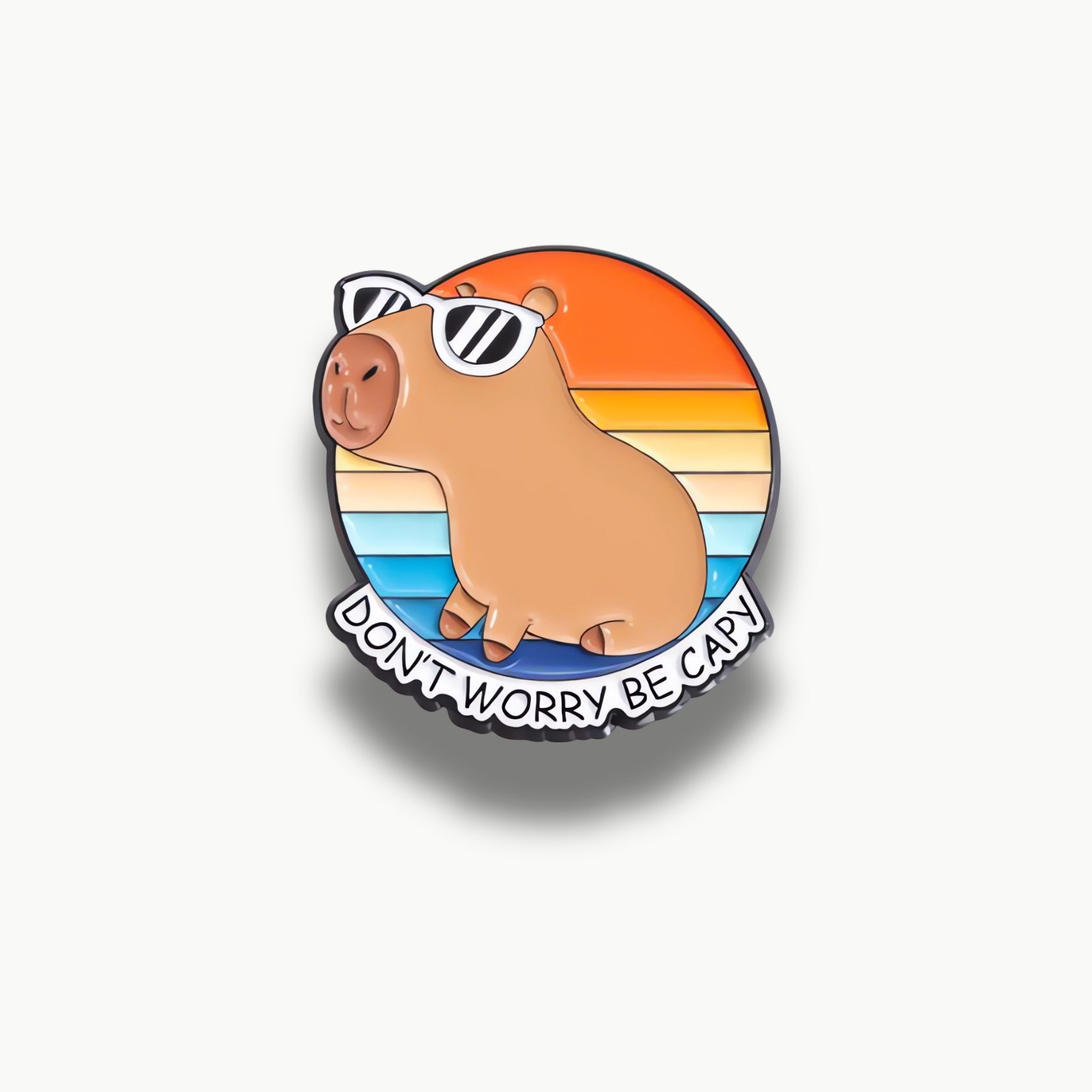 Brošňa s hnedou kapibarou v čiernych okuliaroch na pruhovanom pozadí s oranžovou a modrou farbou. Pod kapibarou je nápis "Don't Worry Be Capy".