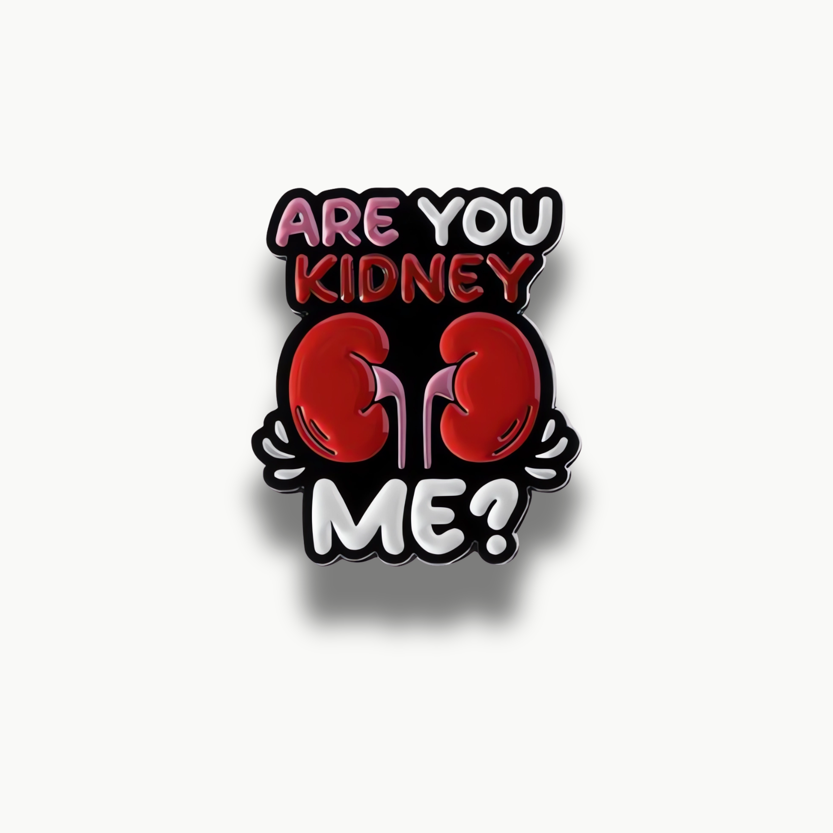 Smaltovaná brošňa s čiernym podkladom, červenými obličkami a farebným nápisom 'ARE YOU KIDNEY ME?'.